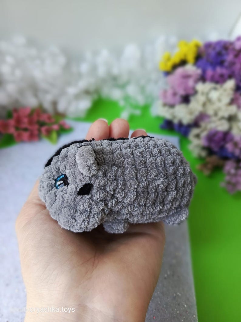 May include: A small, gray plush toy in the shape of a hamster. The toy has black eyes and a black stripe down its back. The hamster is held in a hand, with a blurred background of flowers.