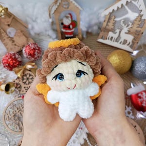 Op de afbeelding: Een handgemaakte, pluche engelpop met een witte body, bruin haar en blauwe ogen. De engel heeft gele vleugels en een halo. De pop wordt in iemands handen gehouden, met kerstversieringen op de achtergrond.