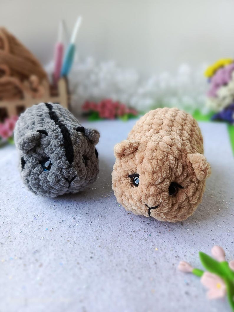 May include: Two handmade crochet guinea pig toys. One is gray with a black stripe, the other is tan. Both have black eyes and small ears. The toys are made of soft, textured yarn and are sitting on a white surface.