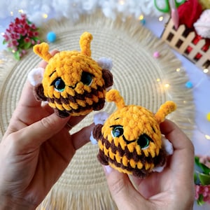 May include: Two handmade, plush bee toys. The bees are yellow and brown with white wings, black eyes, and green eye accents. The toys are held in a person's hands.