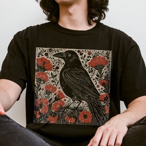 May include: A black t-shirt with a graphic print of a black crow perched on a branch with red flowers. The crow is surrounded by a floral design with leaves and stems.
