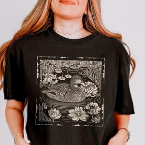 May include: A black t-shirt with a vintage-style illustration of a duck swimming in a pond with lily pads. The illustration is in a cream color and has a distressed look.