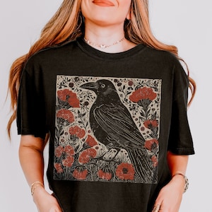 May include: Black t-shirt with a graphic print of a crow perched on a branch with red flowers. The crow is black and the flowers are red and white.