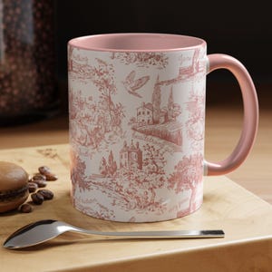 Puede incluir: Taza de cerámica blanca con interior y asa rosas, con un diseño de toile de Jouy rojo que representa escenas con edificios y árboles, y las palabras "headstrong girl". La taza está sobre una tabla de madera con una cuchara y un macaron.