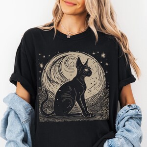 May include: Black t-shirt with a graphic print of a black cat sitting in front of a crescent moon with stars. The moon has a swirling pattern.