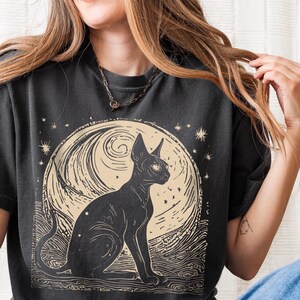 May include: A black t-shirt with a graphic of a black cat sitting in front of a full moon with stars. The moon is a cream color with a swirling pattern.