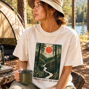 May include: Cream-colored t-shirt with a nature scene graphic featuring a red sun, trees, and a winding river. The text "PROTECT WHAT YOU LOVE" is below the graphic. A person is wearing the shirt and a tan bucket hat, holding a green speckled mug.