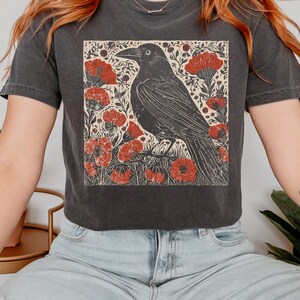 May include: A black t-shirt with a vintage style graphic of a crow in a field of red flowers. The crow is black and the flowers are red with a cream background.