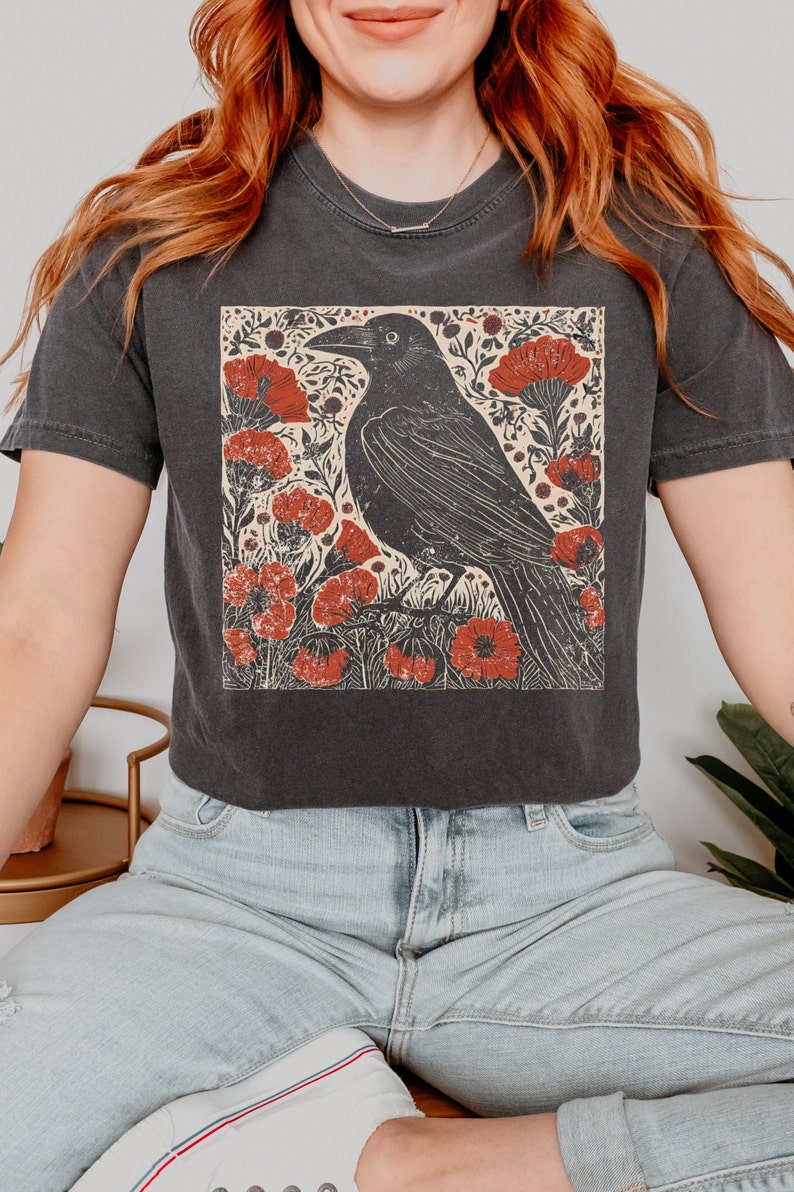 May include: A black t-shirt with a vintage style graphic of a crow in a field of red flowers. The crow is black and the flowers are red with a cream background.