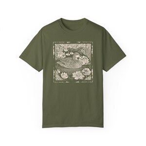May include: A green t-shirt with a white print of a duck swimming in a pond with lily pads. The duck is in the center of the print, and the lily pads are around it. The print is in a rectangular frame.