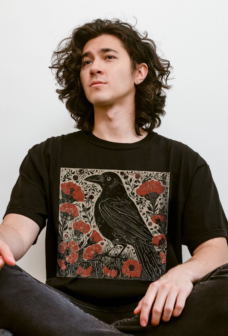 May include: A black t-shirt with a graphic print of a black crow perched on a branch with red flowers. The crow is surrounded by a floral design with leaves and stems.
