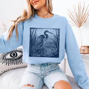 Puede incluir: Sudadera azul claro con una ilustraci&oacute;n azul oscuro de una garza en el agua. La garza est&aacute; representada con l&iacute;neas detalladas, de pie entre juncos. La sudadera es de color azul claro liso.
