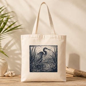 May include: Natural canvas tote bag with a dark blue print of a heron in a pond, surrounded by reeds. The heron is illustrated in a detailed linocut style. The bag has long handles for carrying over the shoulder.