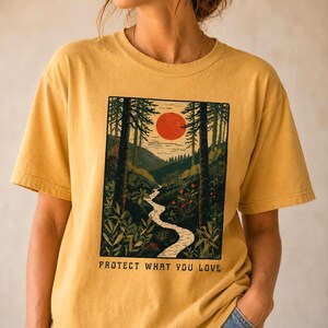 May include: Mustard yellow t-shirt featuring a nature scene with a river, trees, and a red sun. The graphic includes the text "PROTECT WHAT YOU LOVE" in a rectangular frame. The shirt has short sleeves and a relaxed fit.