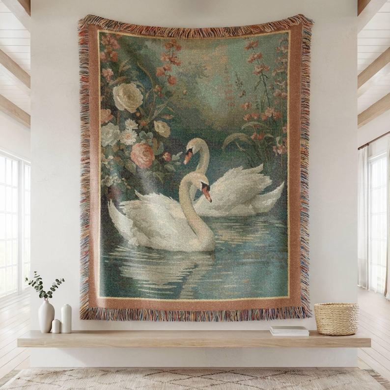 Puede incluir: Un tapiz tejido que representa dos cisnes blancos en un lago, rodeados de flores. El tapiz tiene un borde con flecos en tonos marrones y beige, creando una est&eacute;tica vintage. La obra de arte se exhibe en una pared.