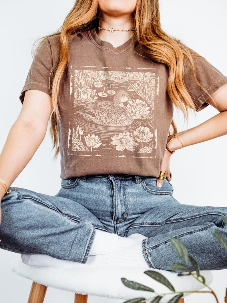 May include: A brown t-shirt with a vintage-style illustration of a duck swimming in a pond with lily pads. The illustration is in a square frame.