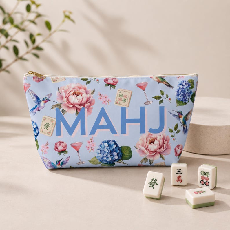 May include: A light blue zippered pouch featuring the text MAHJ in large blue letters, surrounded by a floral pattern of pink peonies, blue hydrangeas, hummingbirds, cocktail glasses, and mahjong tiles, with four loose mahjong tiles nearby.