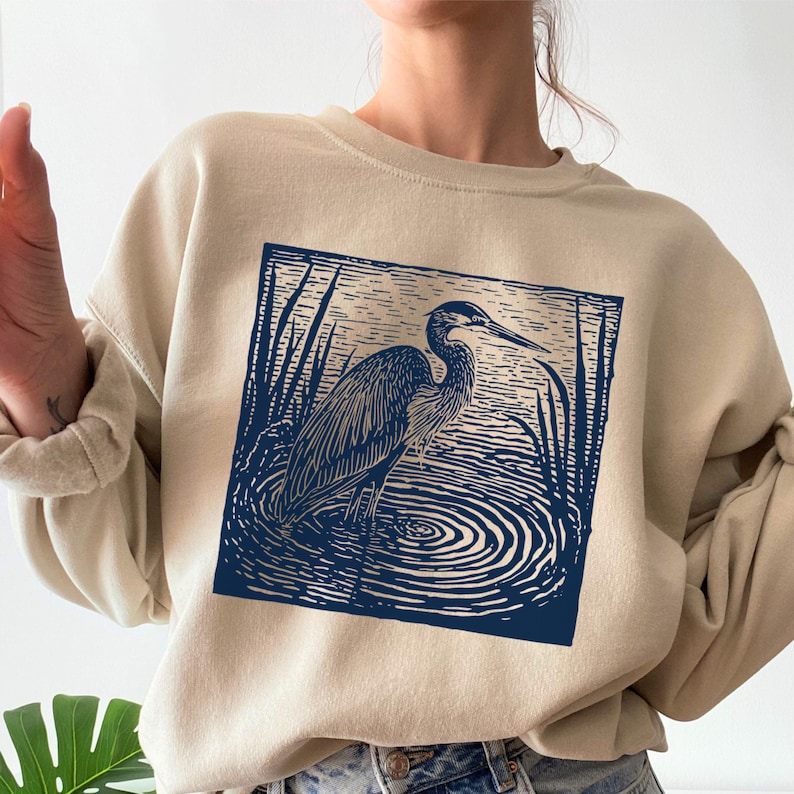 Puede incluir: Sudadera beige con un dise&ntilde;o azul marino de una garza en el agua con juncos. La garza est&aacute; en un marco cuadrado. La sudadera tiene cuello redondo y mangas largas.