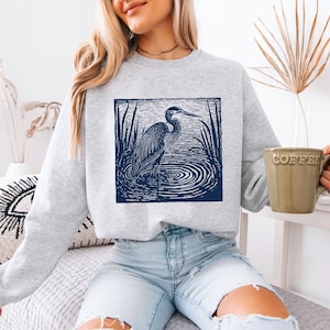 Puede incluir: Una sudadera gris claro con un gr&aacute;fico azul marino de una garza en el agua, rodeada de juncos. La garza est&aacute; en un marco cuadrado. La persona de la imagen lleva vaqueros azul claro y sostiene una taza de caf&eacute; verde.