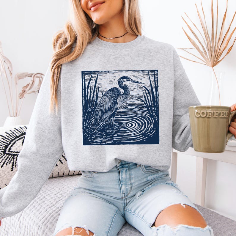 Puede incluir: Una sudadera gris claro con un gr&aacute;fico azul marino de una garza en el agua, rodeada de juncos. La garza est&aacute; en un marco cuadrado. La persona de la imagen lleva vaqueros azul claro y sostiene una taza de caf&eacute; verde.