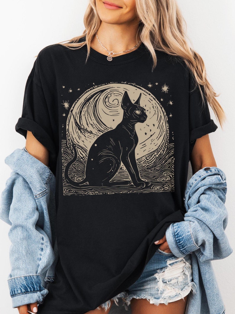 May include: Black t-shirt with a graphic print of a black cat sitting in front of a crescent moon with stars. The moon has a swirling pattern.