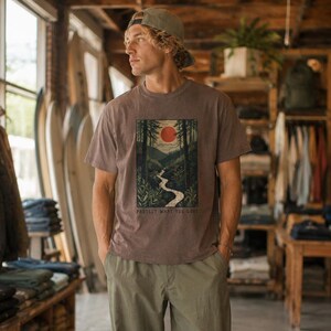 May include: A brown t-shirt with a nature scene graphic featuring a red sun, river, and trees. The text "PROTECT WHAT YOU LOVE" is at the bottom. The person is wearing a green cap and khaki pants.
