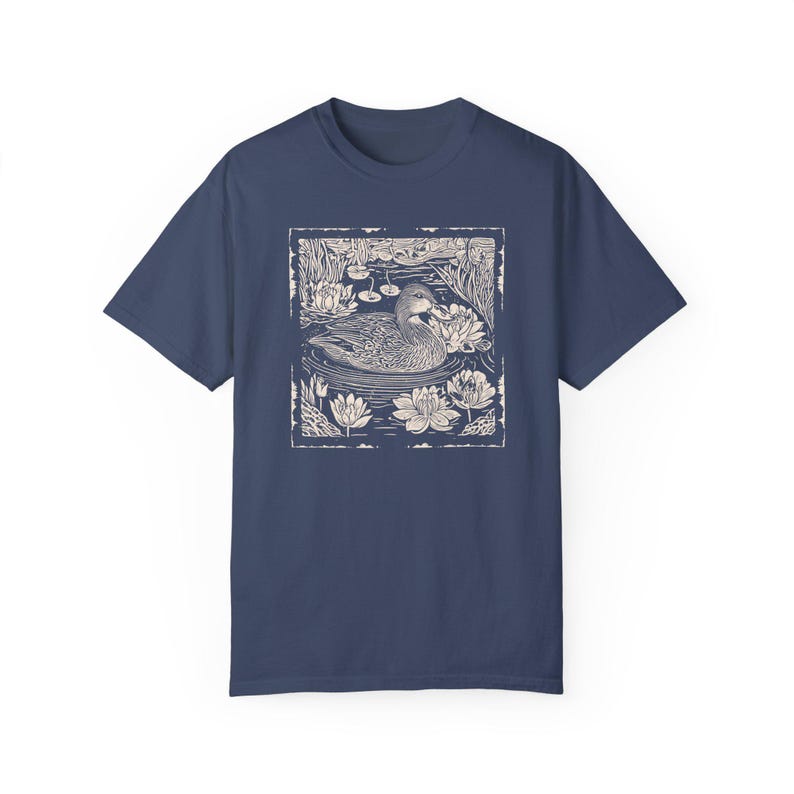 May include: A navy blue t-shirt with a white print of a duck swimming in a pond with lily pads. The print is in a vintage style.