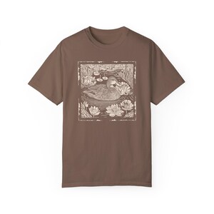 May include: A brown t-shirt with a white print of a duck swimming in a pond with lily pads. The duck is in the center of the print, and the lily pads are around it. The print is framed with a white border.
