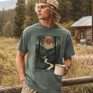 May include: A green t-shirt featuring a landscape design with a red sun, trees, and a winding river. The text "PROTECT WHAT YOU" is printed at the bottom. The person is wearing a tan baseball cap and holding a beige mug.
