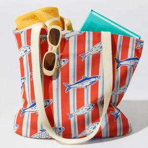 May include: A red and white striped tote bag with a blue fish pattern. The bag has cream-colored handles and contains a yellow towel, sunglasses, and a turquoise book. The bag is designed for carrying beach essentials.