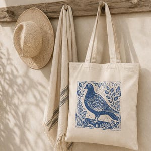 May include: A beige tote bag with a blue pigeon print, a straw hat, and a beige towel hanging on a wooden rack. The tote bag features a blue illustration of a pigeon surrounded by leaves.