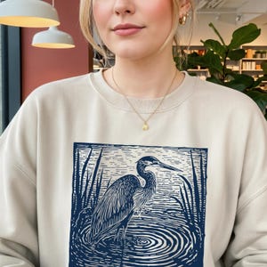Puede incluir: Sudadera beige con un gr&aacute;fico azul y blanco de una garza en el agua. La garza est&aacute; detallada con l&iacute;neas, y el agua tiene c&iacute;rculos conc&eacute;ntricos. La sudadera es de color neutro.