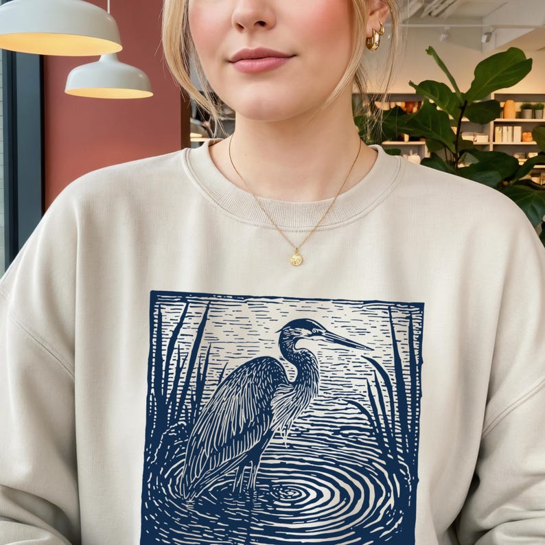 Puede incluir: Sudadera beige con un gr&aacute;fico azul y blanco de una garza en el agua. La garza est&aacute; detallada con l&iacute;neas, y el agua tiene c&iacute;rculos conc&eacute;ntricos. La sudadera es de color neutro.