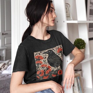 May include: A black t-shirt with a vintage-style illustration of a crow perched on a branch with red flowers. The crow is black and the flowers are a deep red.