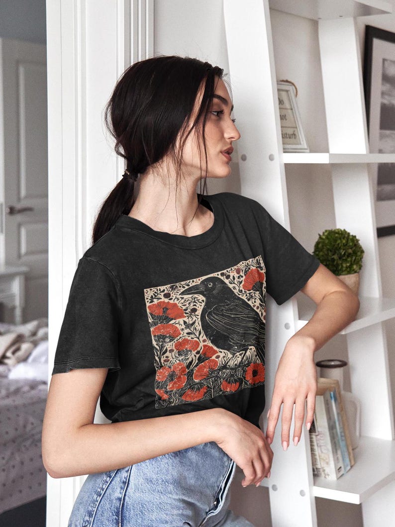 May include: A black t-shirt with a vintage-style illustration of a crow perched on a branch with red flowers. The crow is black and the flowers are a deep red.