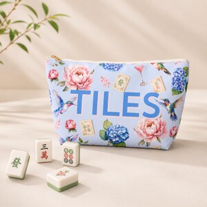 May include: A light blue zippered pouch featuring the word TILES in large blue letters, surrounded by a floral pattern of pink peonies and blue hydrangeas, hummingbirds, and mahjong tiles. Four individual mahjong tiles sit in front of the pouch.