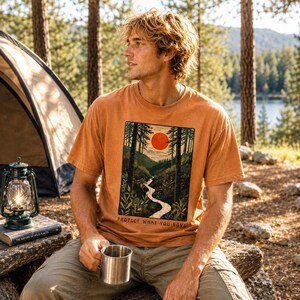 May include: An orange t-shirt with a nature scene graphic and the text "PROTECT WHAT YOU LOVE." The graphic depicts a river winding through a forest with a red sun. The person is sitting outdoors with a metal mug.