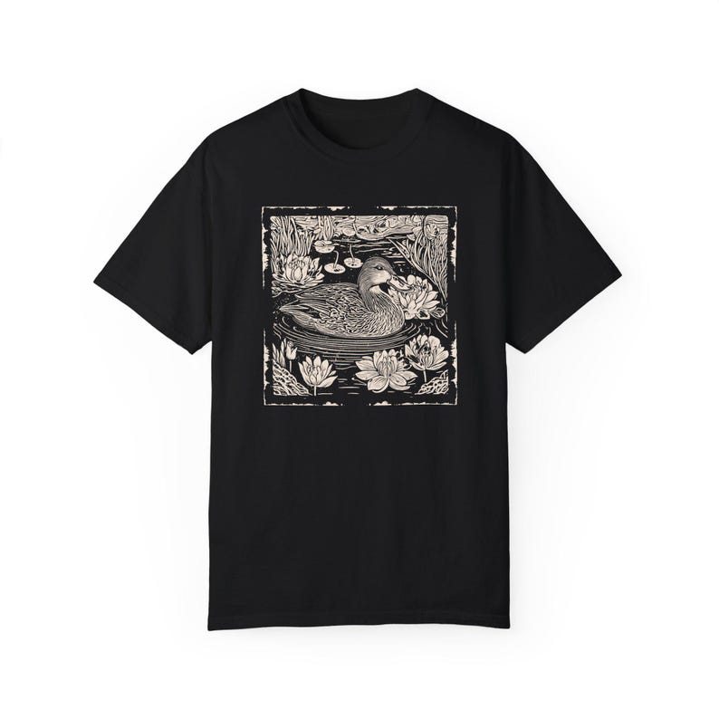 May include: Black t-shirt with a white graphic print of a duck swimming in a pond with lily pads.
