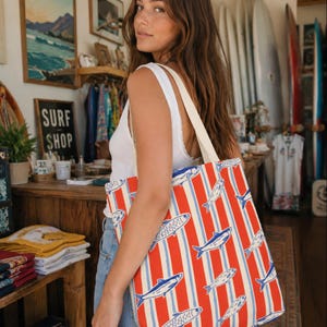 May include: A red and white striped tote bag with a fish pattern. The bag has a cream-colored handle and is being held by a person. The bag's design features blue fish and blue stripes.