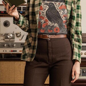 May include: A person wearing a black t-shirt with a graphic print of a black raven and red flowers. The t-shirt is worn under a green and white plaid blazer. The person is wearing brown pants and a brown belt.