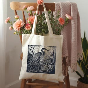May include: A cream tote bag with a blue and white heron illustration. The bag is displayed on a wooden chair, adorned with peach-coloured flowers and a pink blanket. The heron is depicted in water.