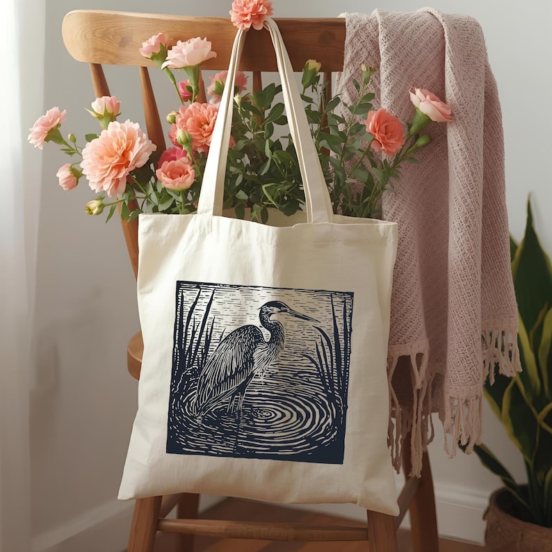 May include: A cream tote bag with a blue and white heron illustration. The bag is displayed on a wooden chair, adorned with peach-coloured flowers and a pink blanket. The heron is depicted in water.