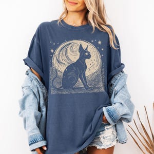 May include: A navy blue t-shirt with a graphic of a black cat sitting in front of a crescent moon with stars. The cat has large, round eyes and a long, thin tail.
