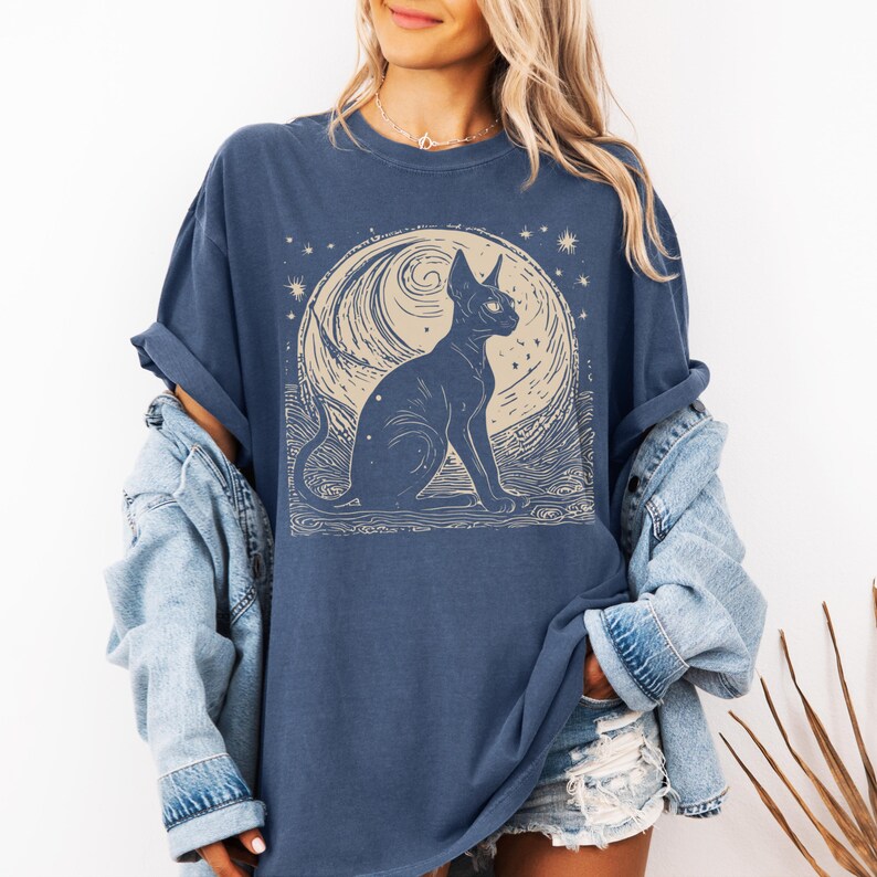 May include: A navy blue t-shirt with a graphic of a black cat sitting in front of a crescent moon with stars. The cat has large, round eyes and a long, thin tail.