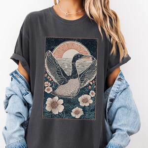 May include: A dark gray t-shirt with a vintage-style graphic of a bird flying in front of a sunset. The graphic is surrounded by flowers and has a distressed look.