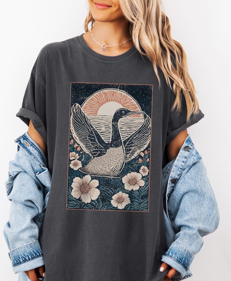 May include: A dark gray t-shirt with a vintage-style graphic of a bird flying in front of a sunset. The graphic is surrounded by flowers and has a distressed look.