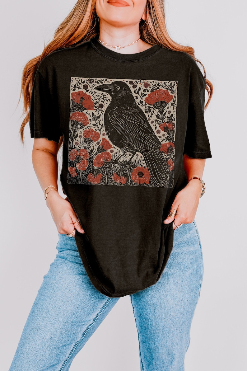 May include: Black t-shirt with a graphic print of a crow perched on a branch with red flowers. The crow is black and the flowers are red and white.