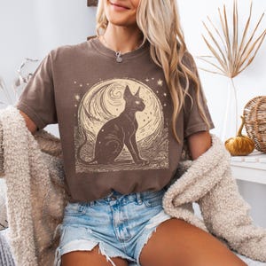 May include: A brown t-shirt with a graphic of a black cat sitting in front of a full moon with stars. The moon has a swirling pattern and the cat has large, round eyes.