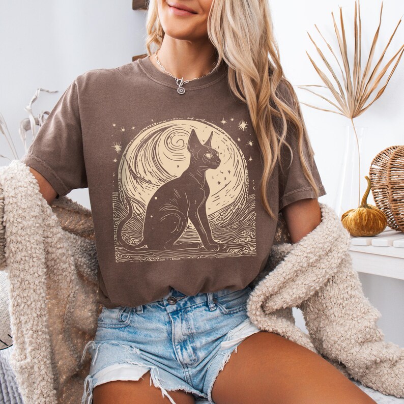 May include: A brown t-shirt with a graphic of a black cat sitting in front of a full moon with stars. The moon has a swirling pattern and the cat has large, round eyes.