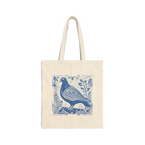 May include: Cream tote bag with a blue linocut print of a pigeon on a branch, framed by leaves. The bag has two handles and is made of a durable fabric. Ideal for carrying shopping or other everyday items.